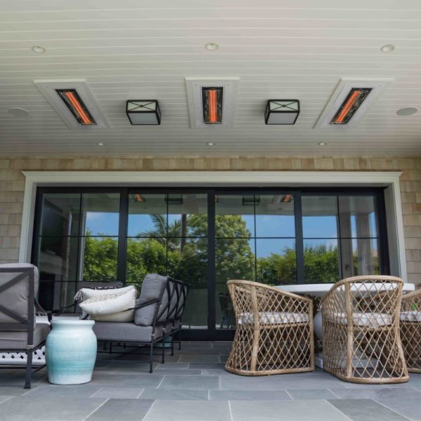 Modern patio area featuring stylish outdoor furniture, including a gray sofa and woven chairs, with a dining table and decorative vase. The ceiling is equipped with infrared heaters and pendant lights, with large glass doors providing a view of lush greenery outside.