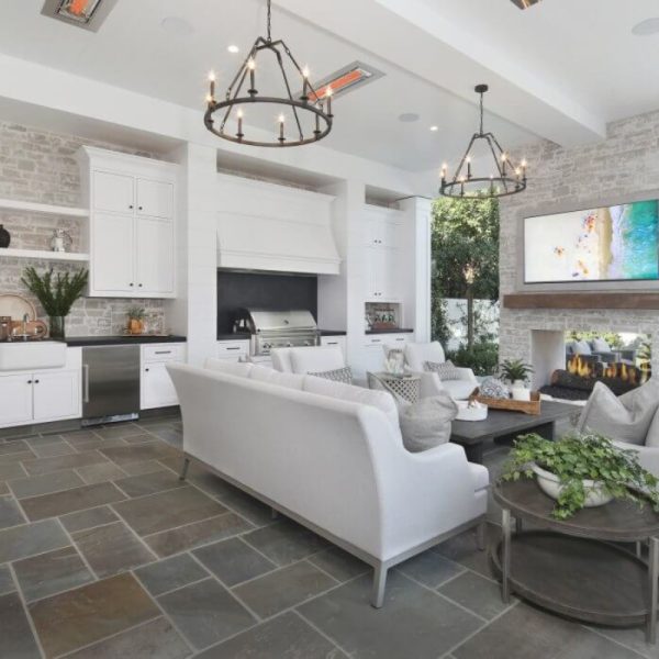 Modern living room with a white sofa, stone fireplace, and a large TV, featuring a kitchen area with stainless steel appliances and open shelving. Bright and airy design with natural light and greenery.