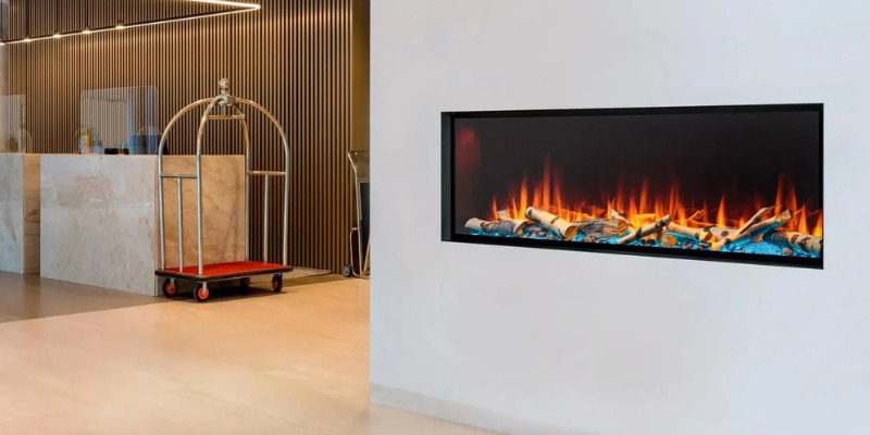 Modern electric fireplace featuring realistic flames and logs, set against a sleek wall in a contemporary lobby with a reception desk and luggage cart in the background.